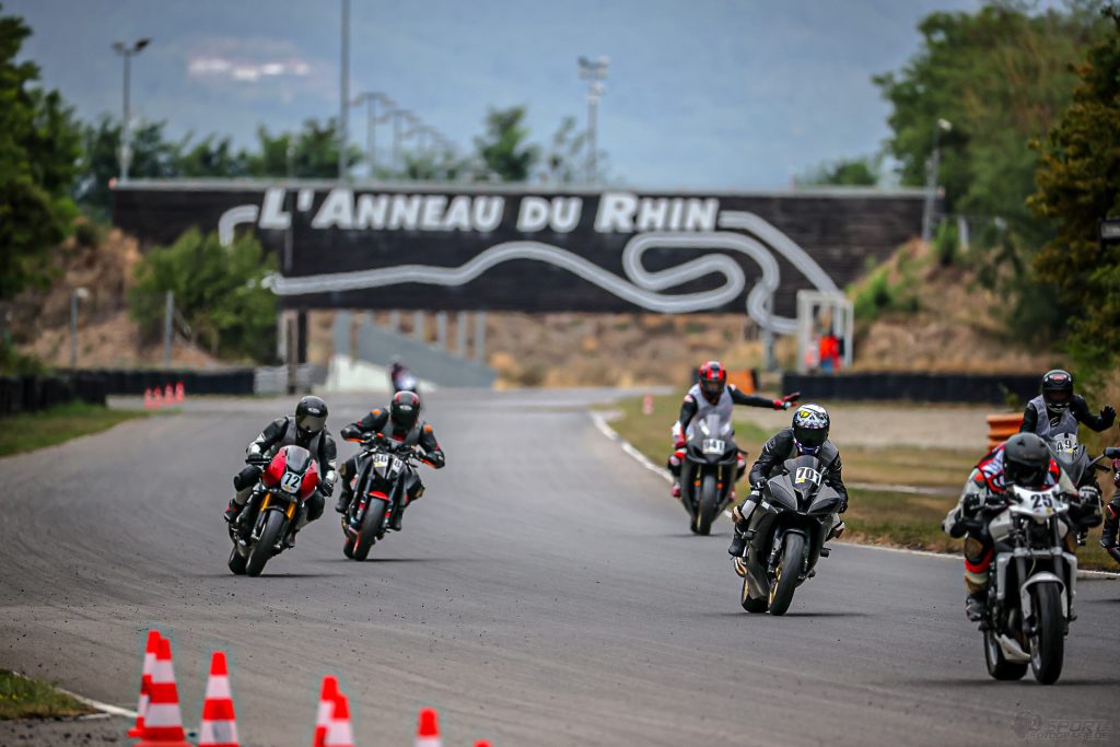 Teilnehmende der instruktionsgeführten Gruppen sind mit ihren Motorrädern auf der Rennstrecke unterwegs. Im Hintergrund sieht man den großen Anneau du Rhin Bogen mit Schrifzug.