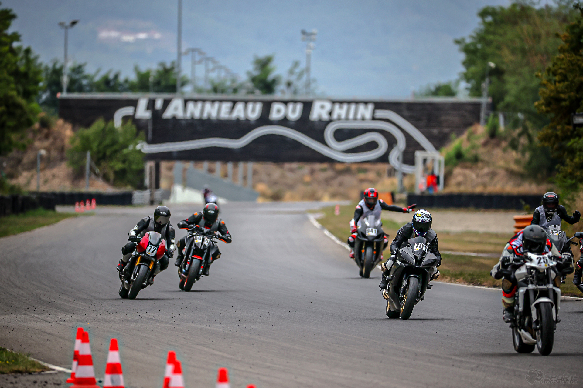 Teilnehmende der instruktionsgeführten Gruppen sind mit ihren Motorrädern auf der Rennstrecke unterwegs. Im Hintergrund sieht man den großen Anneau du Rhin Bogen mit Schrifzug.